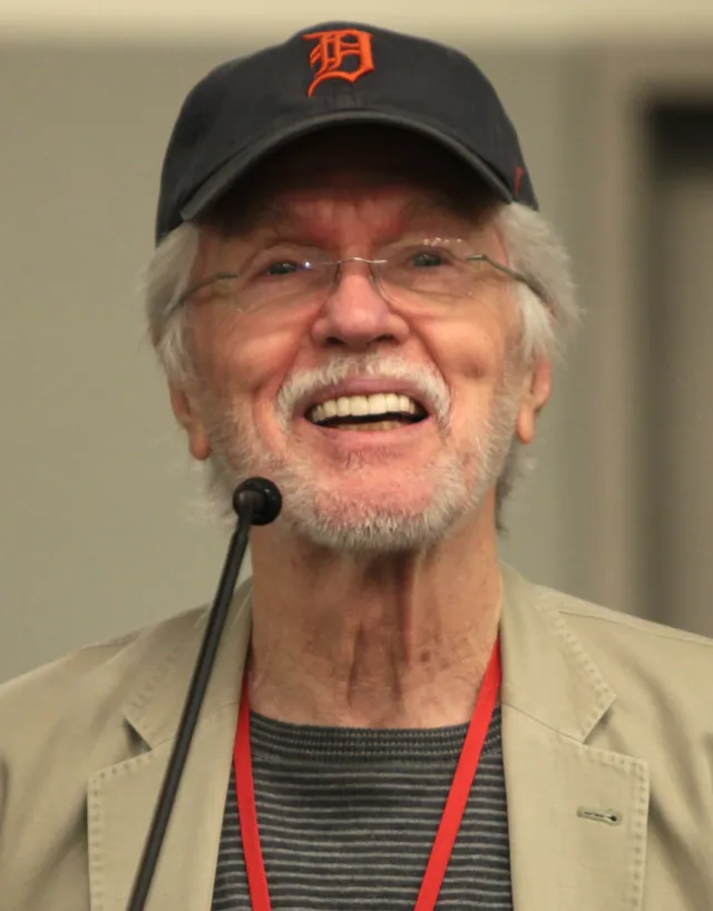 Tom Skerritt laughing at a microphone, wearing glasses and a Detroit Tigers cap