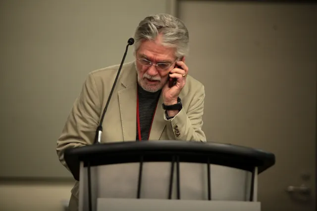 Tom Skerritt at a lectern, one hand raised to the side of his head