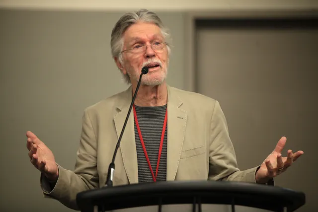 Tom Skerritt speaking at a lectern with both hands spread open