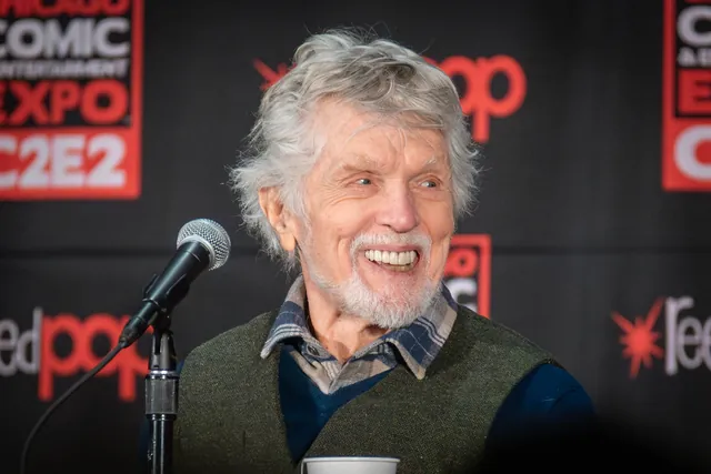 Tom Skerritt, white-haired and bearded, smiling broadly beside a microphone